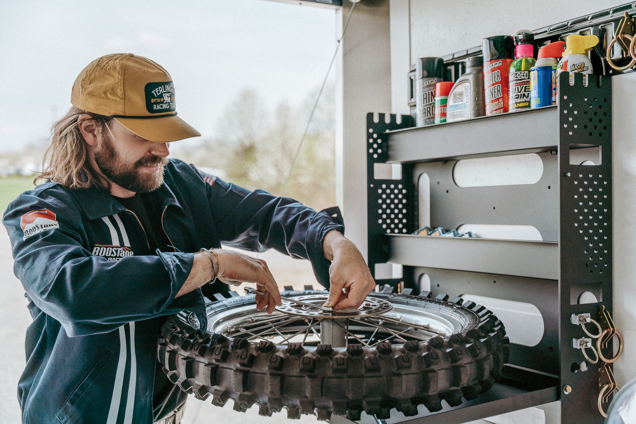 Dirt bike trailer wall organizer with open shelf supporting a dirt bike tire while it is being repaired with a wrench. The organizer is also storing several lubricants and chemicals that might be needed.