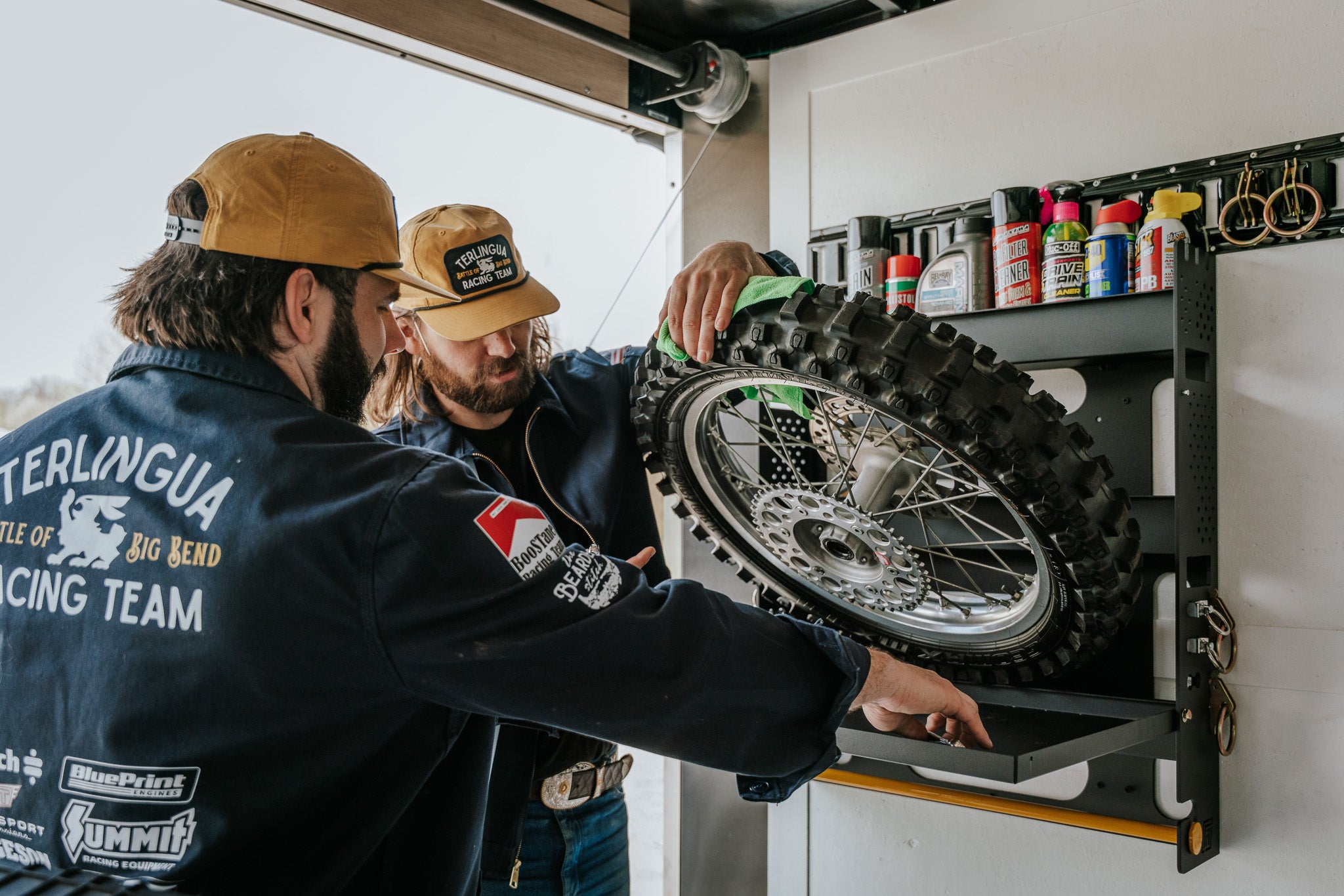 Two men placing a motorcycle tire on the open shelf of the MotoProHQ Dirt Bike Trailer Wall Organizer. There are various lubricants and chemicals stored on the organizer shelf as well as storage for max or e-track tie down rings.