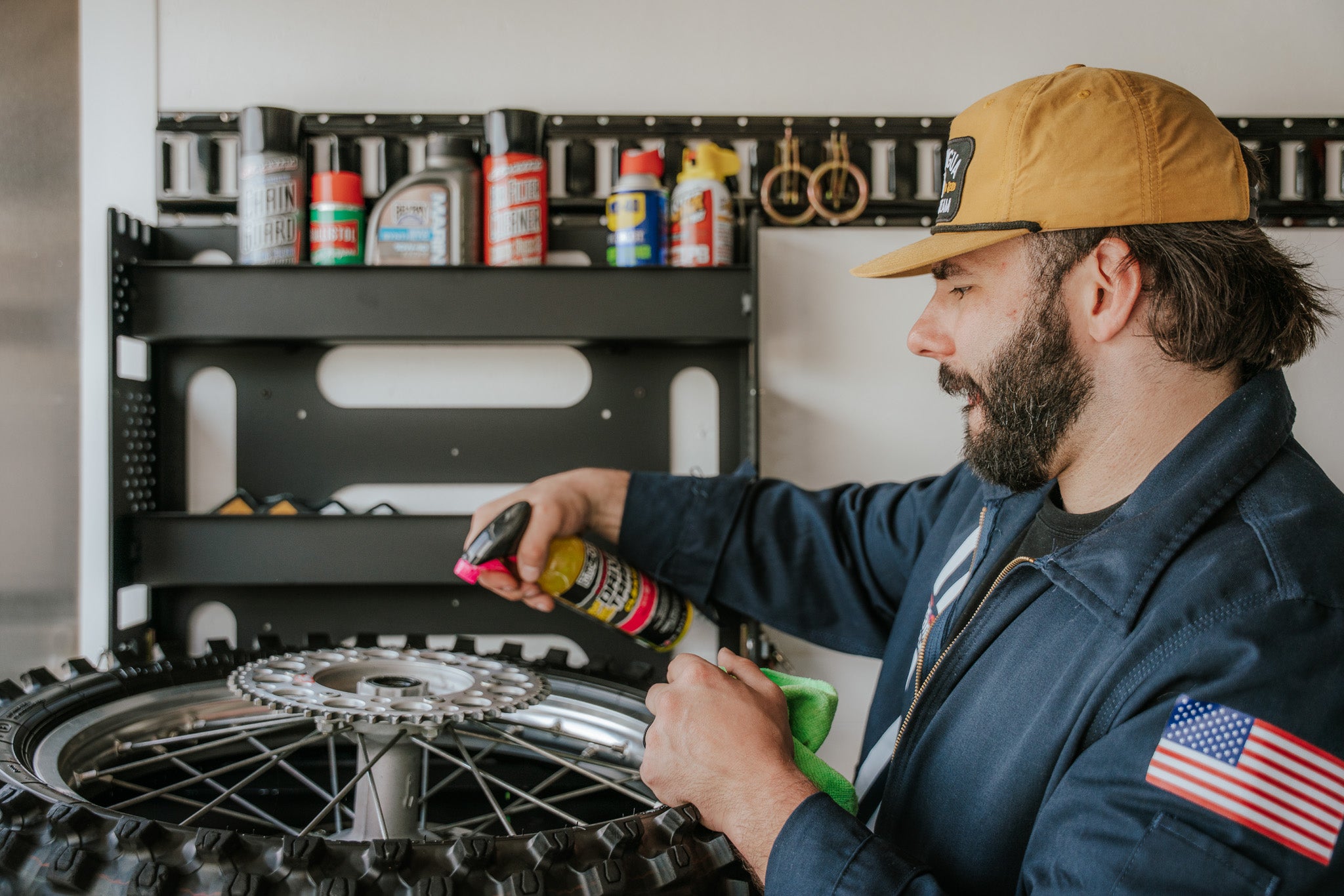 Man cleaning a motorcycle tire with tools and supplies in the background. The tire is resting on the opened shelf of the MotoProHQ Dirt Bike Trailer Wall Organizer.
